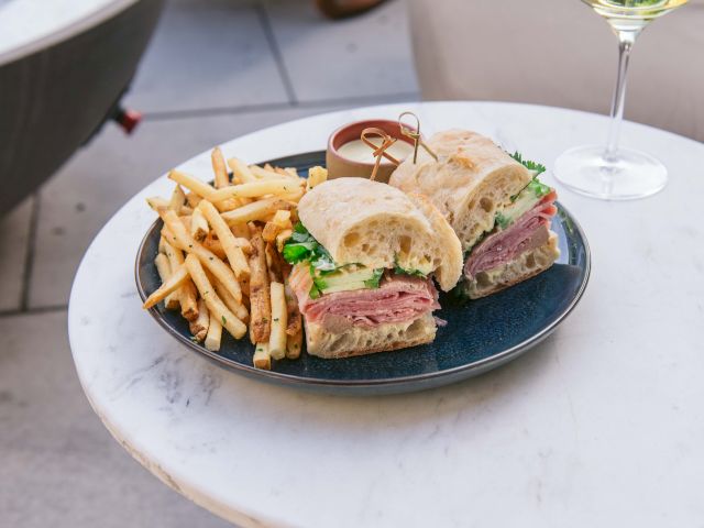 A plate with a sandwich, fries, and a dipping sauce on a table, next to a glass of white wine.