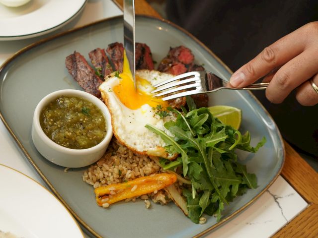 A person cuts into a sunny-side-up egg on steak, served with rice, arugula, carrots, sauce, and lime on a plate.