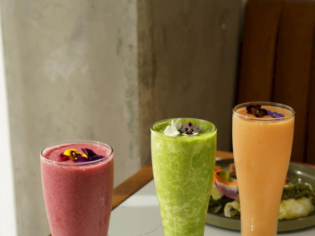 Three tall glasses of smoothies—red, green, and orange—each garnished with an edible flower, on a table next to a wooden booth.