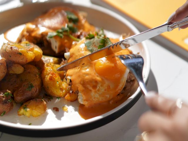 The image shows a plate with eggs, potatoes, and garnish, being cut with a knife and fork. A drink and book are in the background.