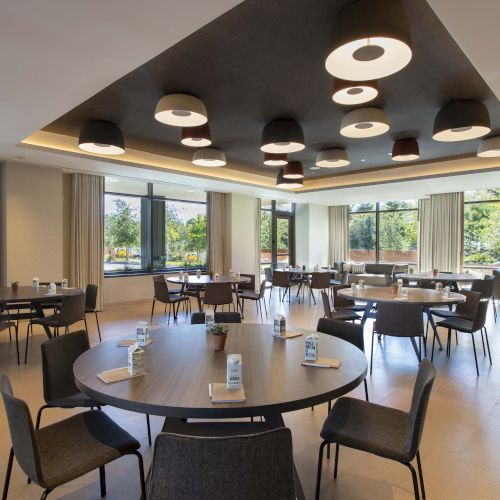 A modern, well-lit meeting room with round tables, chairs, and large windows showing greenery outside.