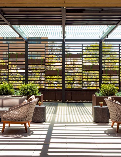 Outdoor seating area with modern furniture, plants, and slatted overhead structure allowing light to filter through, creating shadows.