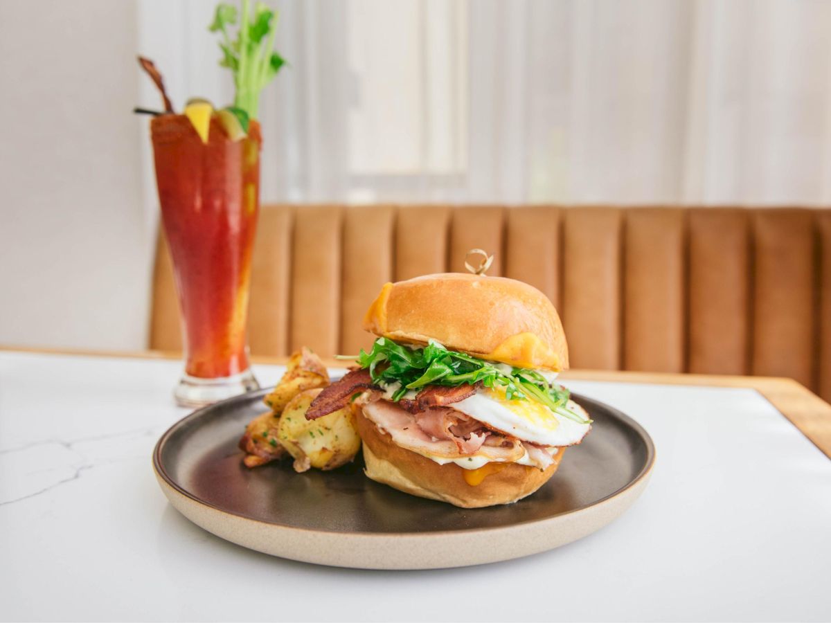 A sandwich with greens on a plate, accompanied by a drink and a side dish on a table.