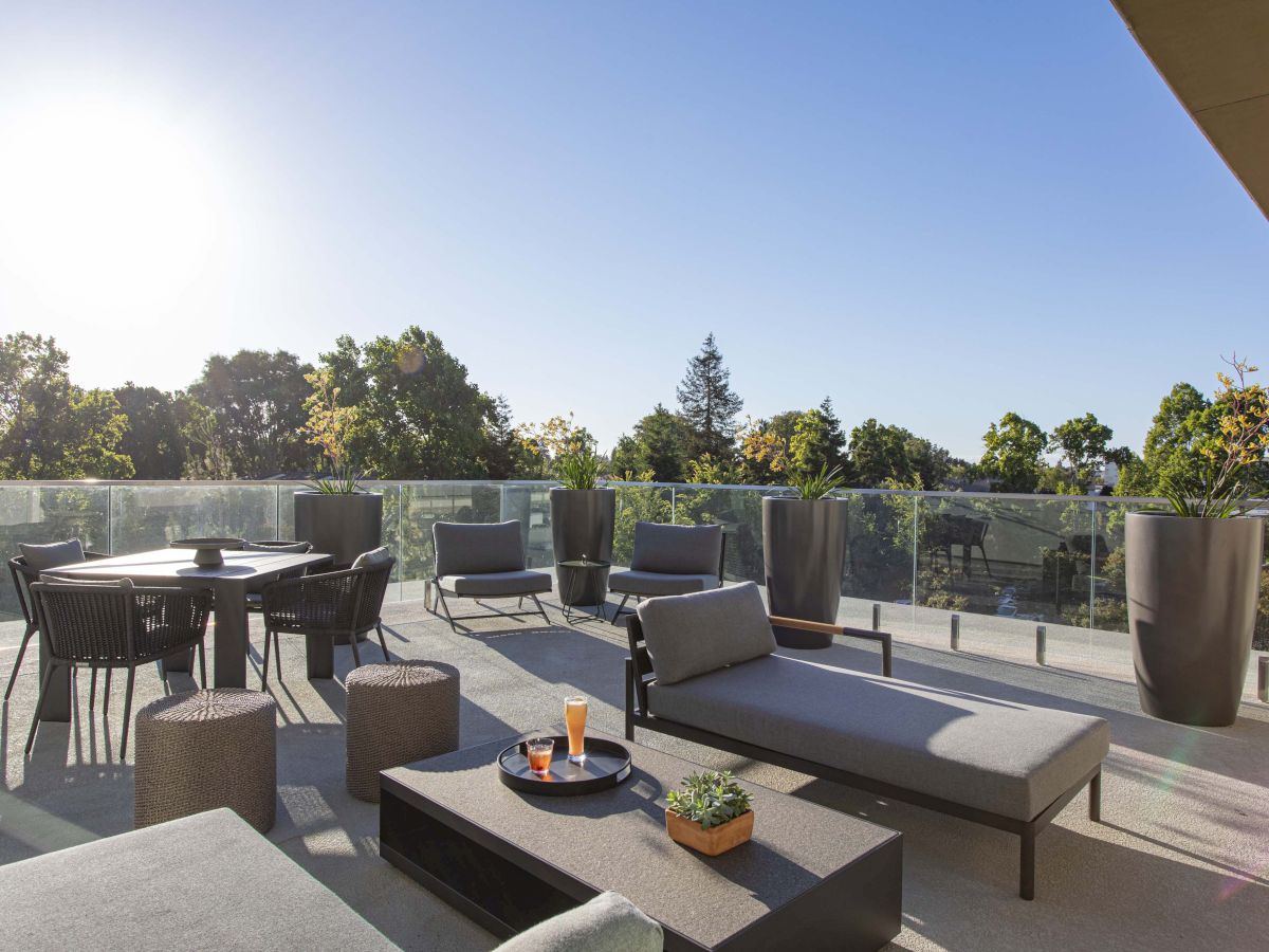 A rooftop patio with lounge chairs, a table, potted plants, drinks on a tray, and a clear view of the sky and trees beyond.