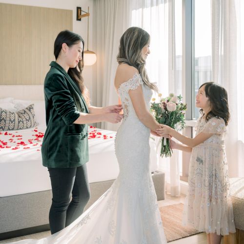 A bride in a white dress holds a bouquet, assisted by two women, with red petals on a bed, in a sunlit room.
