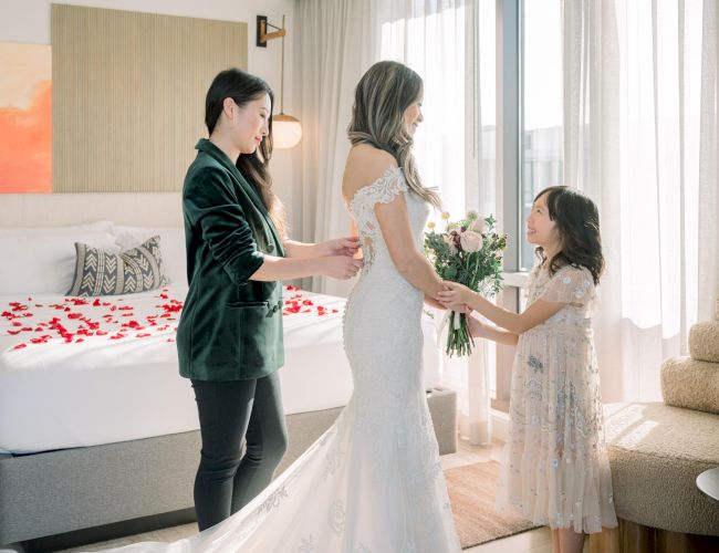 A bride in a white dress holds a bouquet, assisted by two women, with red petals on a bed, in a sunlit room.