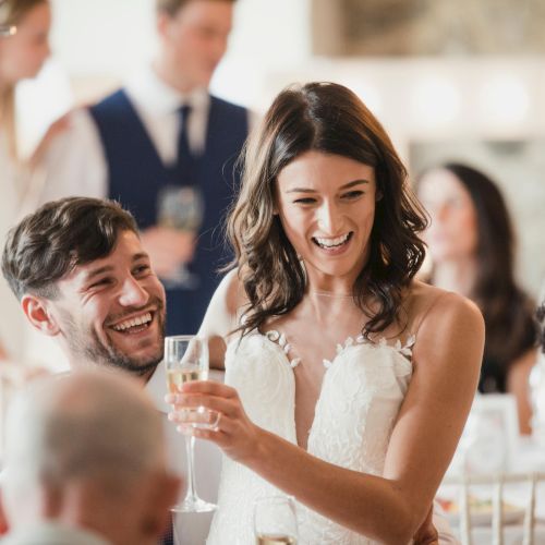 A happy bride in a wedding dress holds a champagne glass, surrounded by laughing guests at a celebration.