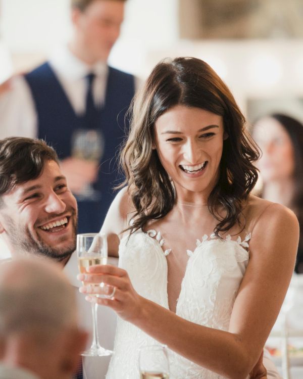 A happy bride in a wedding dress holds a champagne glass, surrounded by laughing guests at a celebration.
