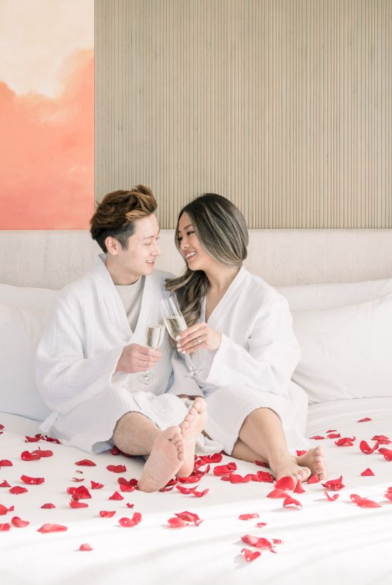 A couple in robes sits on a bed covered with rose petals, holding champagne glasses, with a pink painting and bedside lamps in the room.