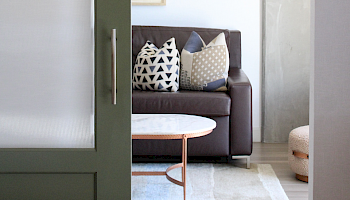 A dark open door reveals a modern living room with a brown sofa, patterned cushions, and a small round coffee table.