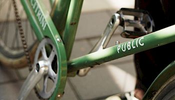 A close-up of a green bicycle with &ldquo;PUBLIC&rdquo; on the frame, parked on a stone sidewalk.
