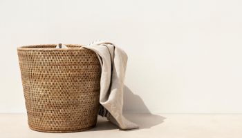 A woven basket with a light-colored cloth draped over it stands against a plain wall on a flat surface.
