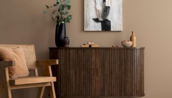 A cozy room with a wooden chair, a sideboard, a rug, and a vase with leaves. Abstract art hangs above on a beige wall.