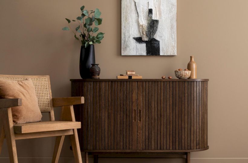 A cozy room with a wooden chair, a sideboard, a rug, and a vase with leaves. Abstract art hangs above on a beige wall.
