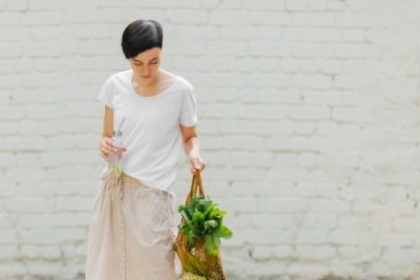 A person in a white shirt holds a bag with greens and a bottle, standing against a white brick wall.