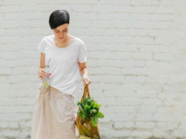 A person in a white shirt holds a bag with greens and a bottle, standing against a white brick wall.