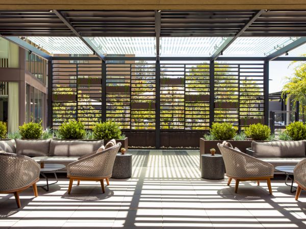 Modern outdoor patio with cushioned sofas, woven chairs, tiled floor, potted plants, and a slatted pergola wall.