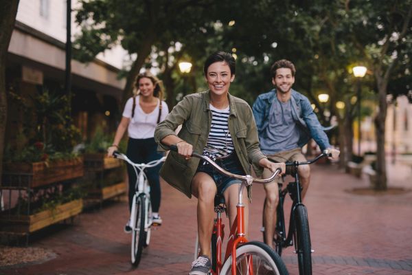 Three people are joyfully riding bicycles down a tree-lined brick path in the evening, smiling as they enjoy the ride together.