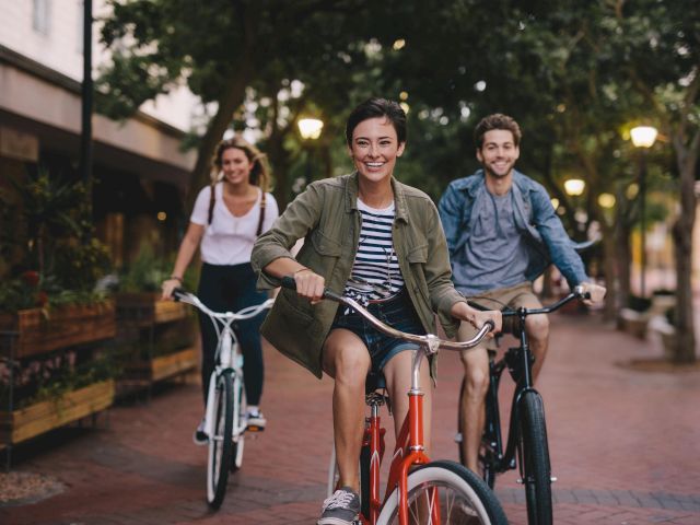 Three people are joyfully riding bicycles on a brick pathway lined with trees and streetlights, enjoying a leisurely outdoor activity.