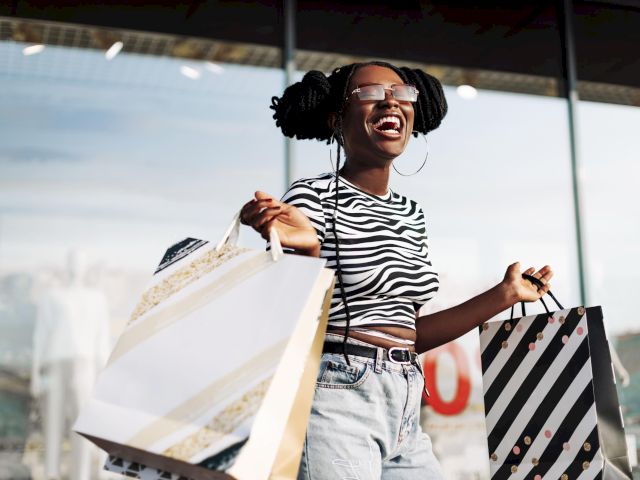 A person joyfully holding shopping bags stands outside a store, wearing a striped top, denim jeans, and sunglasses.