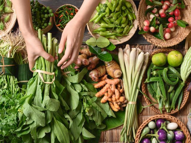 A variety of fresh vegetables, including greens, herbs, and root vegetables, are displayed with hands organizing them.