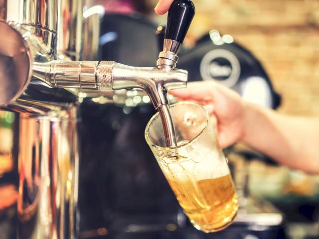 A hand is pouring beer from a tap into a tilted glass, with a blurred background of what looks like a bar or pub setting.