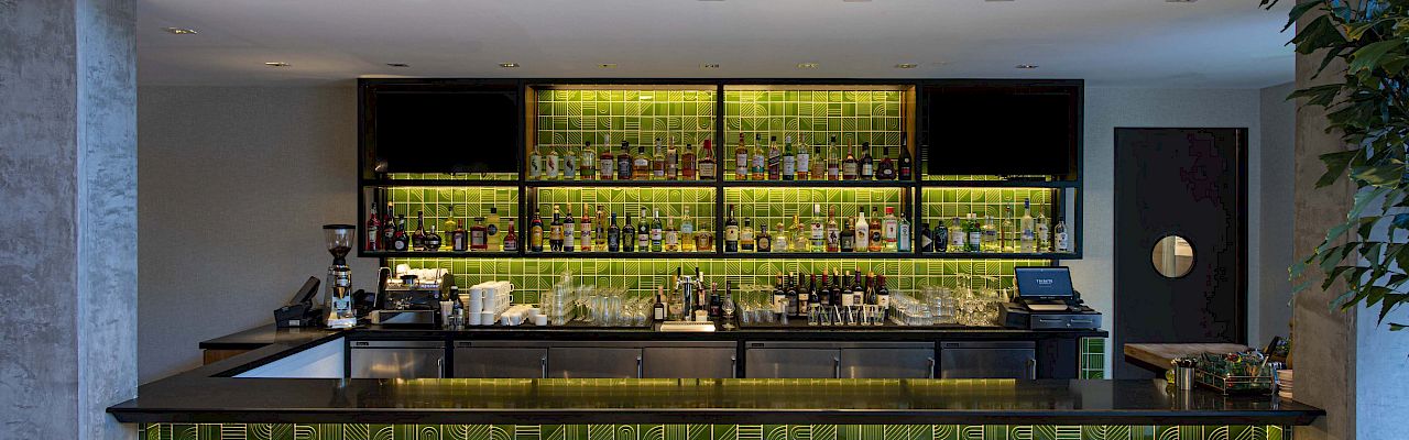 A modern bar with green tile accents, stocked with bottles and glasses, a black countertop, and wooden stools, featuring ambient lighting.