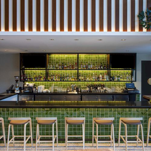 A modern bar with green tile accents, stocked with bottles and glasses, a black countertop, and wooden stools, featuring ambient lighting.