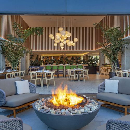 A modern outdoor seating area with a central fire pit, leading into a stylish open dining area with hanging lights and plants.