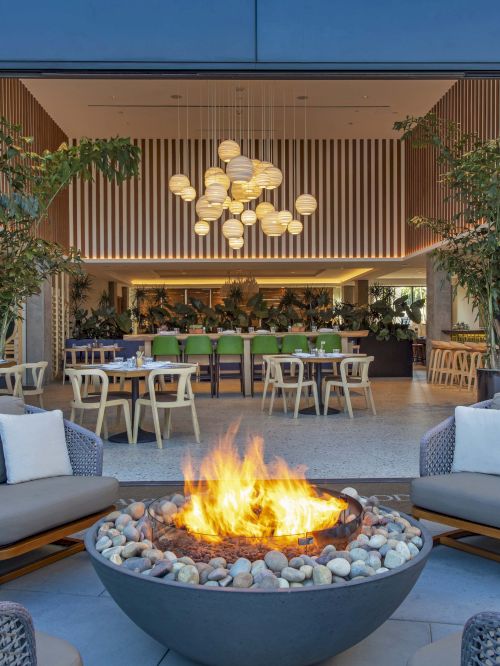 A modern outdoor seating area with a central fire pit, leading into a stylish open dining area with hanging lights and plants.