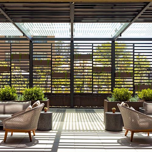 An outdoor patio with cushioned lounge chairs, a tiled floor, a pergola ceiling, and lush potted plants in front of a slatted wall.