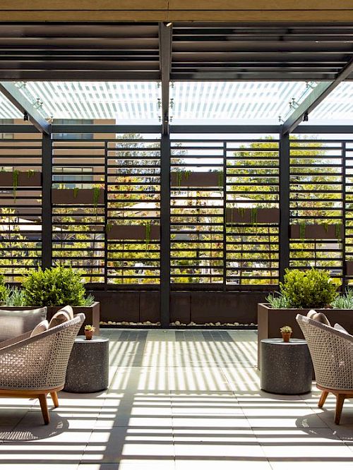 An outdoor patio with cushioned lounge chairs, a tiled floor, a pergola ceiling, and lush potted plants in front of a slatted wall.