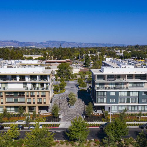 Two modern hotel buildings are adjacent, surrounded by trees, with mountains in the distant background under a clear blue sky.