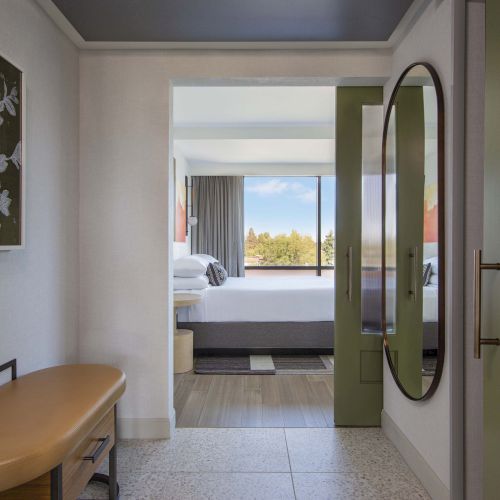 A modern hotel room entrance with a bench, artwork, a mirror, and a view into a bedroom with a large window.