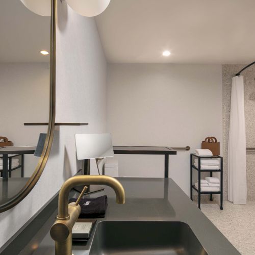 Modern bathroom with a sleek sink, brass faucet, round mirror, shower with curtain, and neatly stacked towels on a shelf.