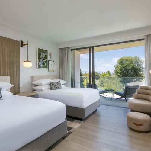 A modern hotel room with two beds, balcony with chairs, and a scenic view. Warm lighting and contemporary decor enhance the interior.
