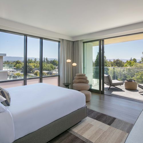 A modern bedroom with large windows, a comfy bed, stylish chair, balcony chairs, and a view of greenery outside.