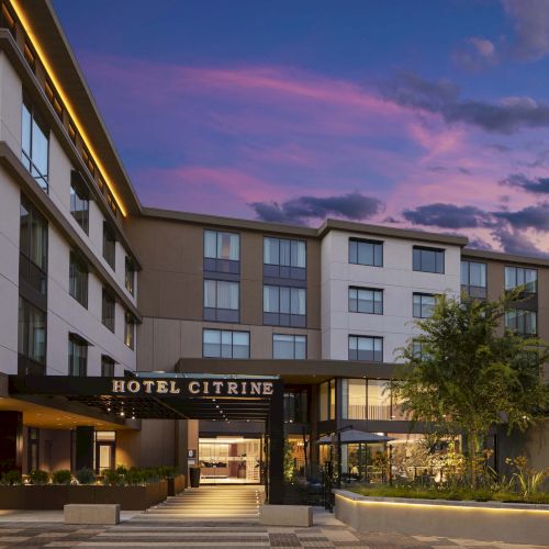 The image shows the exterior of Hotel Citrine with a modern facade, lit signage, and outdoor landscaping under a colorful evening sky.