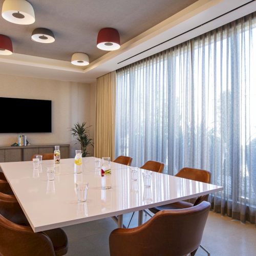 A modern conference room with a white table, leather chairs, a wall-mounted TV, decorative plants, and soft lighting under sheer curtains.