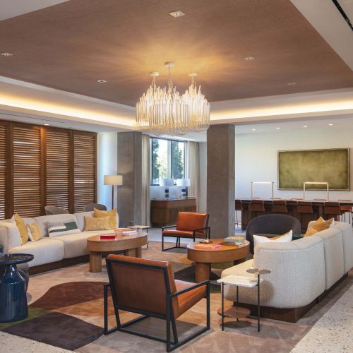 A modern living space with a chandelier, sofas, armchairs, wooden table, and a large plant, leading to a dining area with a long table.