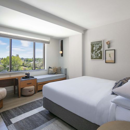 The image shows a modern, minimalistic hotel room with a large window, bed, desk, and stylish decor.