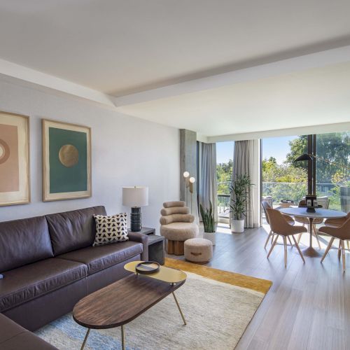Modern living room with a brown sofa, abstract art, round dining table, chairs, plants, and large windows showcasing outdoor greenery.