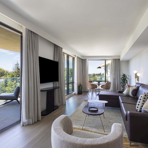 A modern living room with a large sectional, artwork, and a balcony view. There are chairs outside and a round table inside the corner.