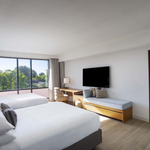 A modern hotel room with two beds, large windows, TV, desk, and a connected living area with sofas and more windows.