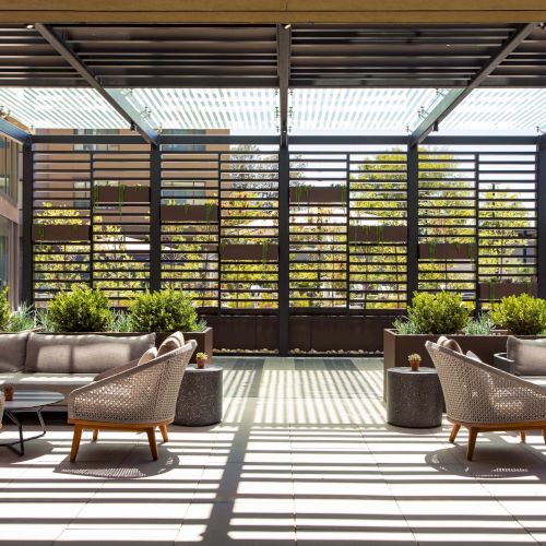 Outdoor seating area with modern wicker furniture, potted plants, and a slatted pergola casting shadows on the tiled floor.