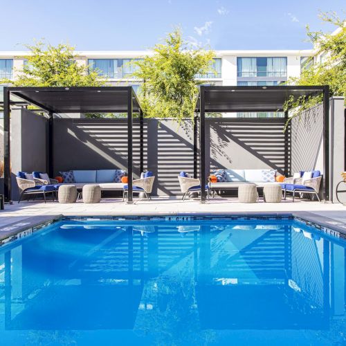 A modern outdoor pool area with cabanas, lounge seating, and greenery, set against the backdrop of a building.