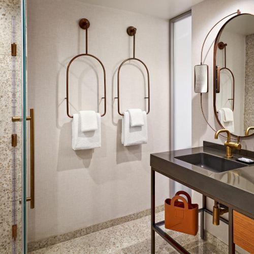 A modern bathroom features a glass shower, towel hangers, a mirror, a black countertop with an oval sink, and a bag underneath.
