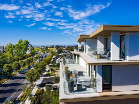 A modern apartment building with glass balconies overlooks a tree-lined street under a blue sky with scattered clouds.