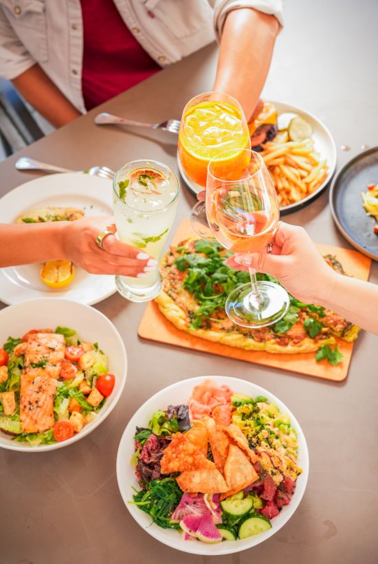 Friends at a table clink glasses over a spread of fresh salads, pizza, and sliced fruit, sharing a celebratory meal together.