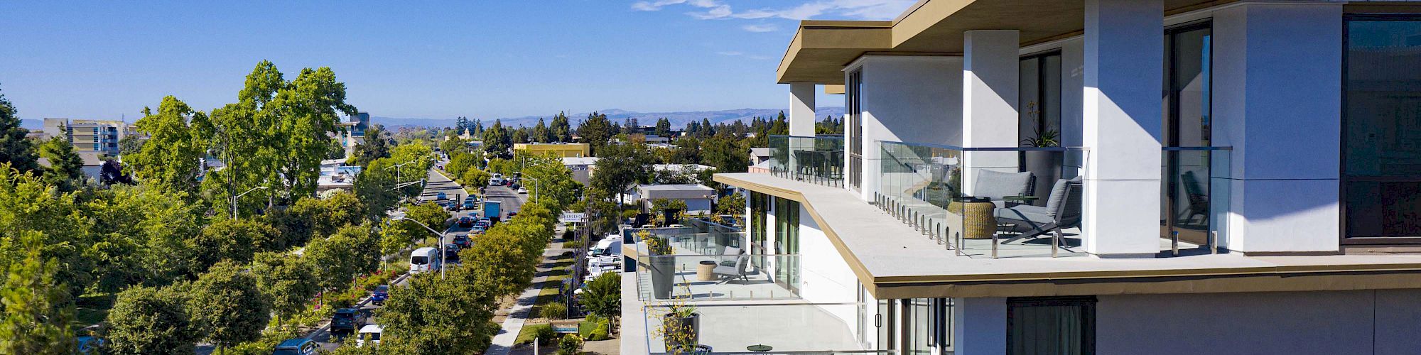 A modern multi-story building with spacious balconies overlooks a tree-lined street, under a bright blue sky with scattered clouds.